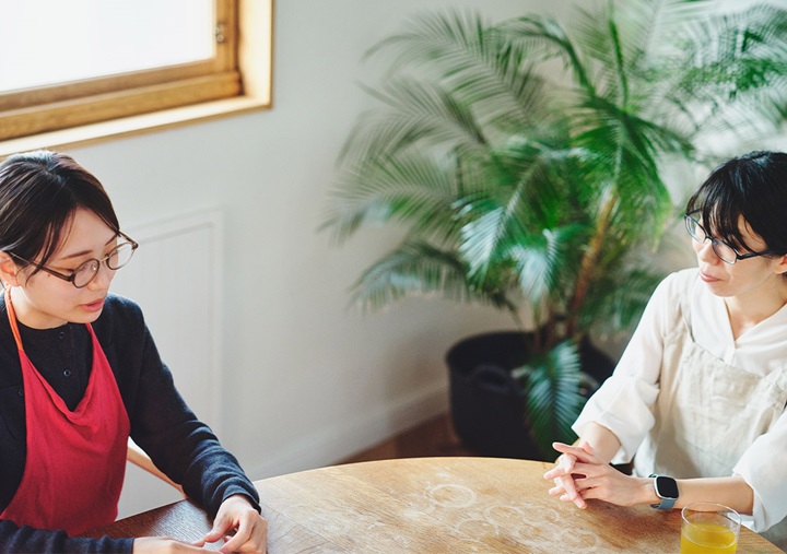Two people talking around a round wooden table, with green houseplants in the background. Akari Hasegawa is on the left, and Ai Sato is on the right.