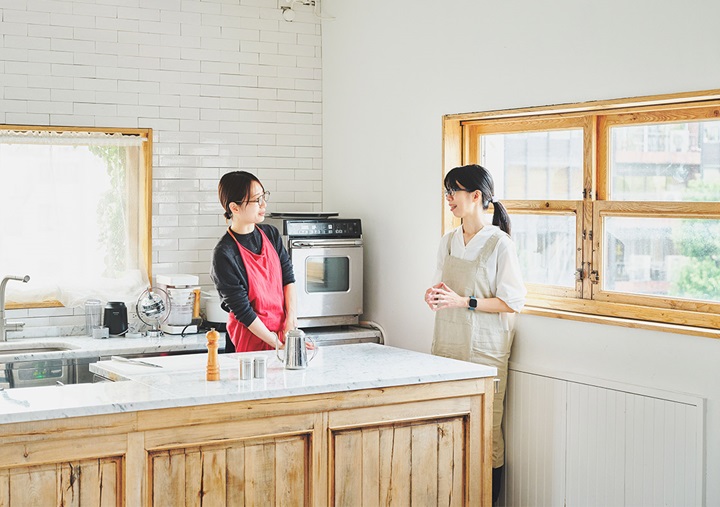 Akari Hasegawa and Ai Sato standing and talking in a sunlit kitchen.