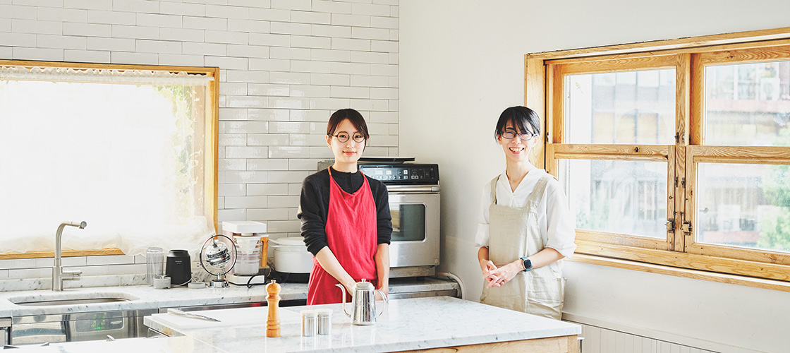 Two people smiling in a sunlit kitchen. Akari Hasegawa is on the left, and Ai Sato is on the right.
